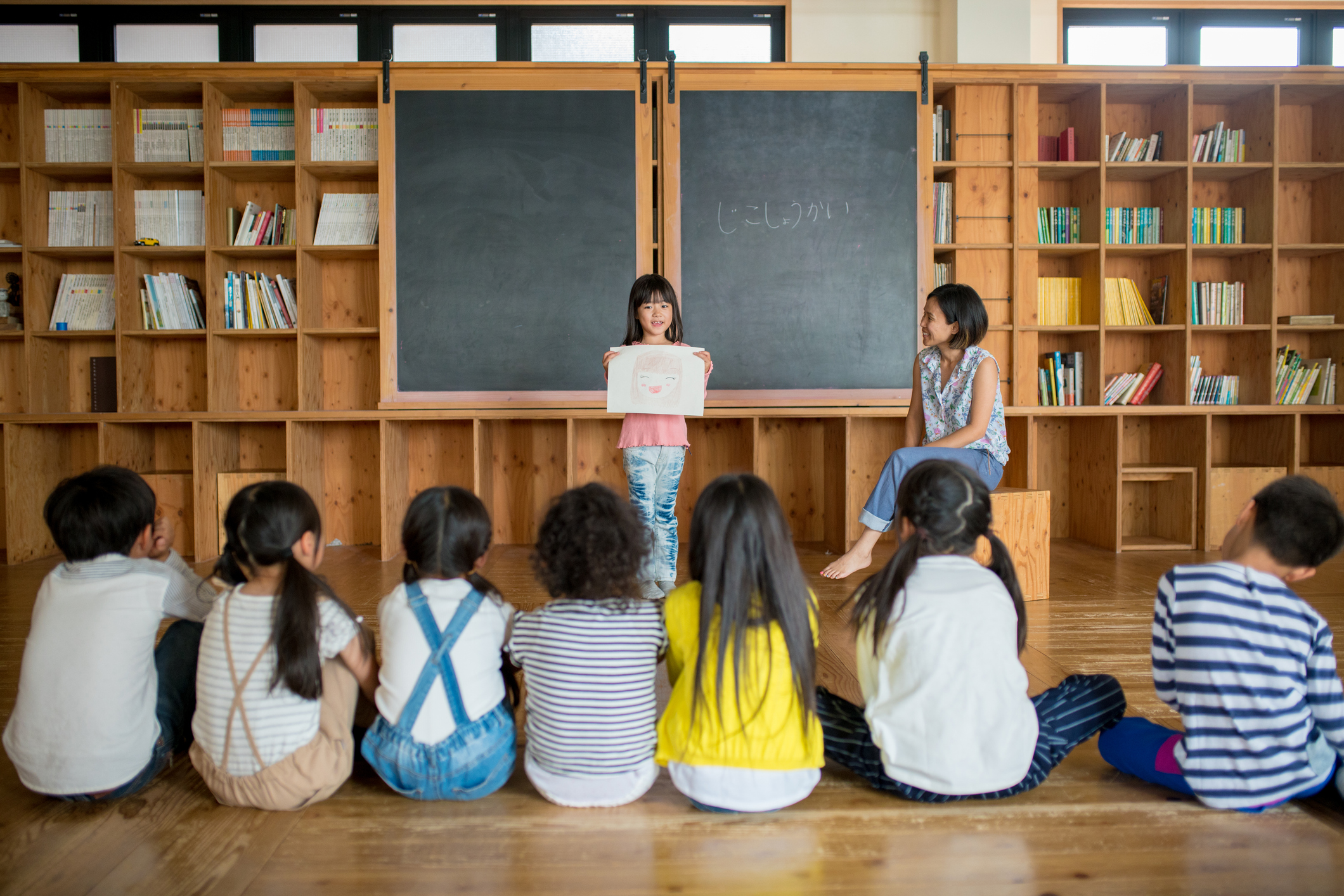 A child stands at the front of a classroom holding up a drawing while classmates sit on the floor and watch.