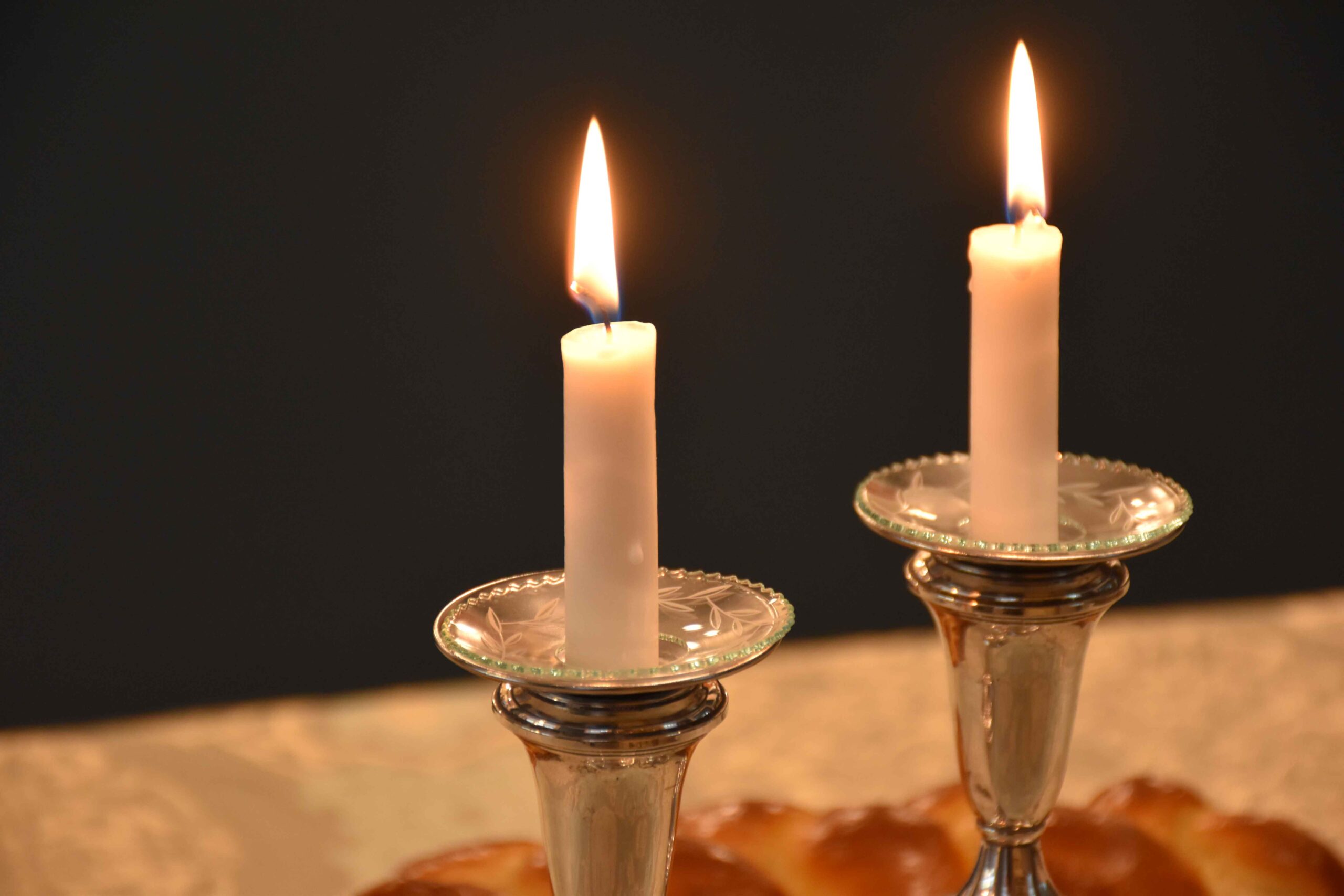 Two Shabbat candles burning brightly in elegant silver holders against a dark background.