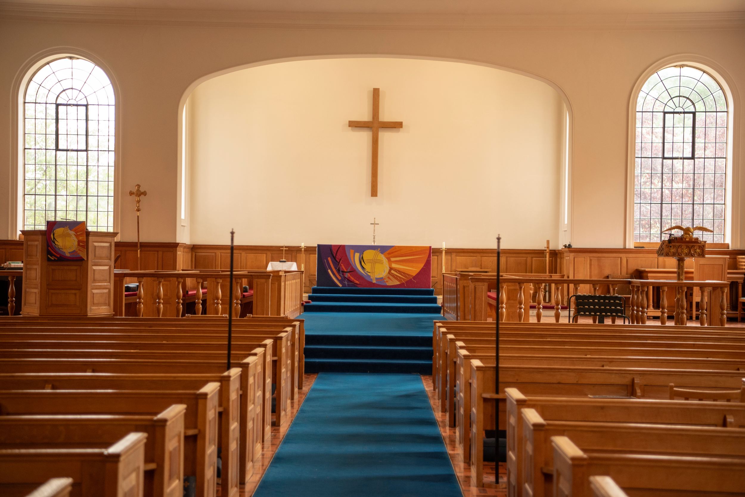 The inside of a church with wooden pews, a blue carpeted aisle, and a large cross at the front above a colourful altar cloth.