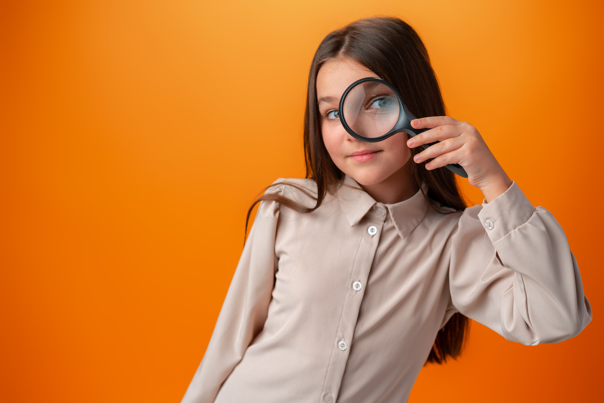 A girl looking through a magnifying glass on an orange background in a studio.