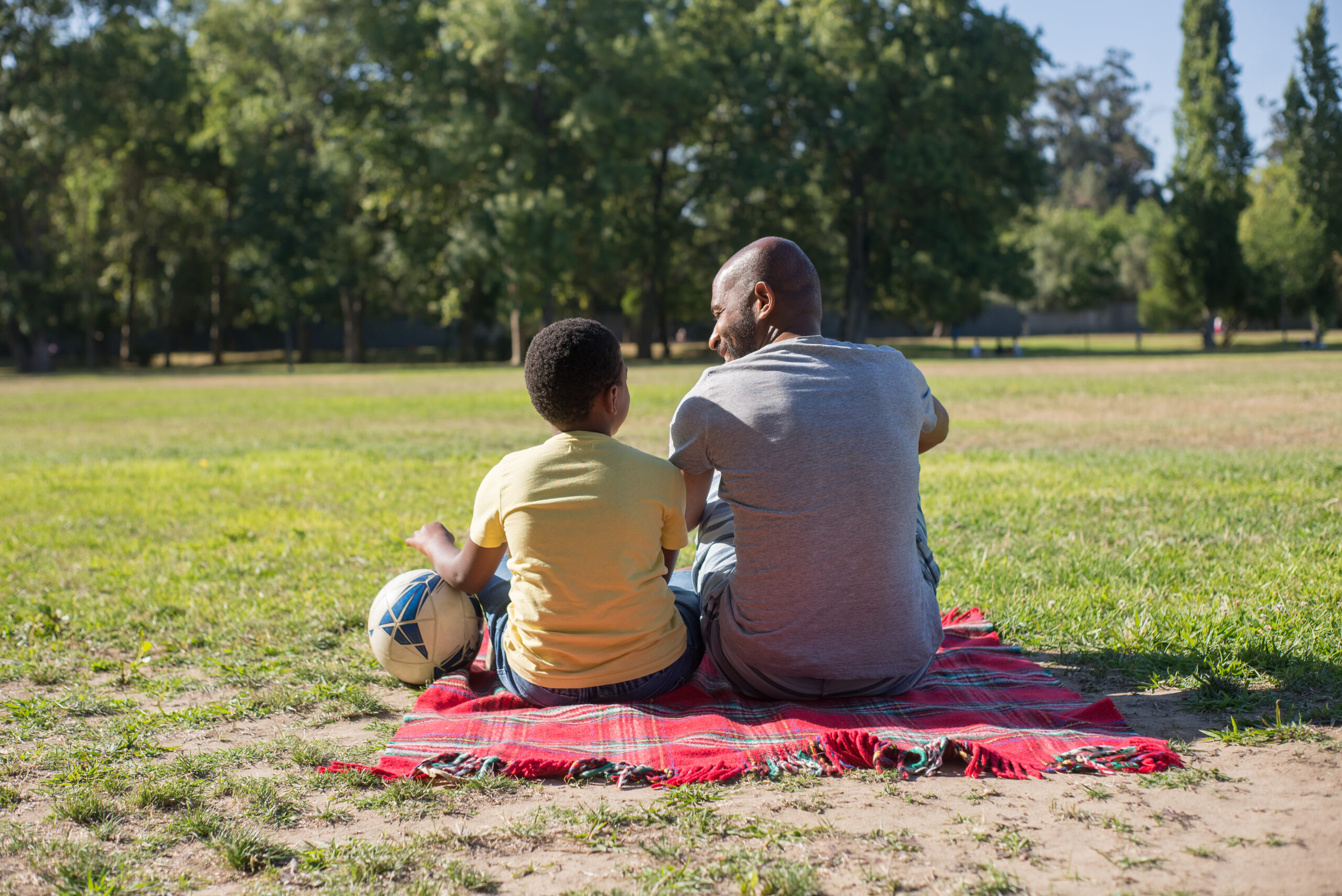 Man and boy sitting on a picnic blanket in a park with a football