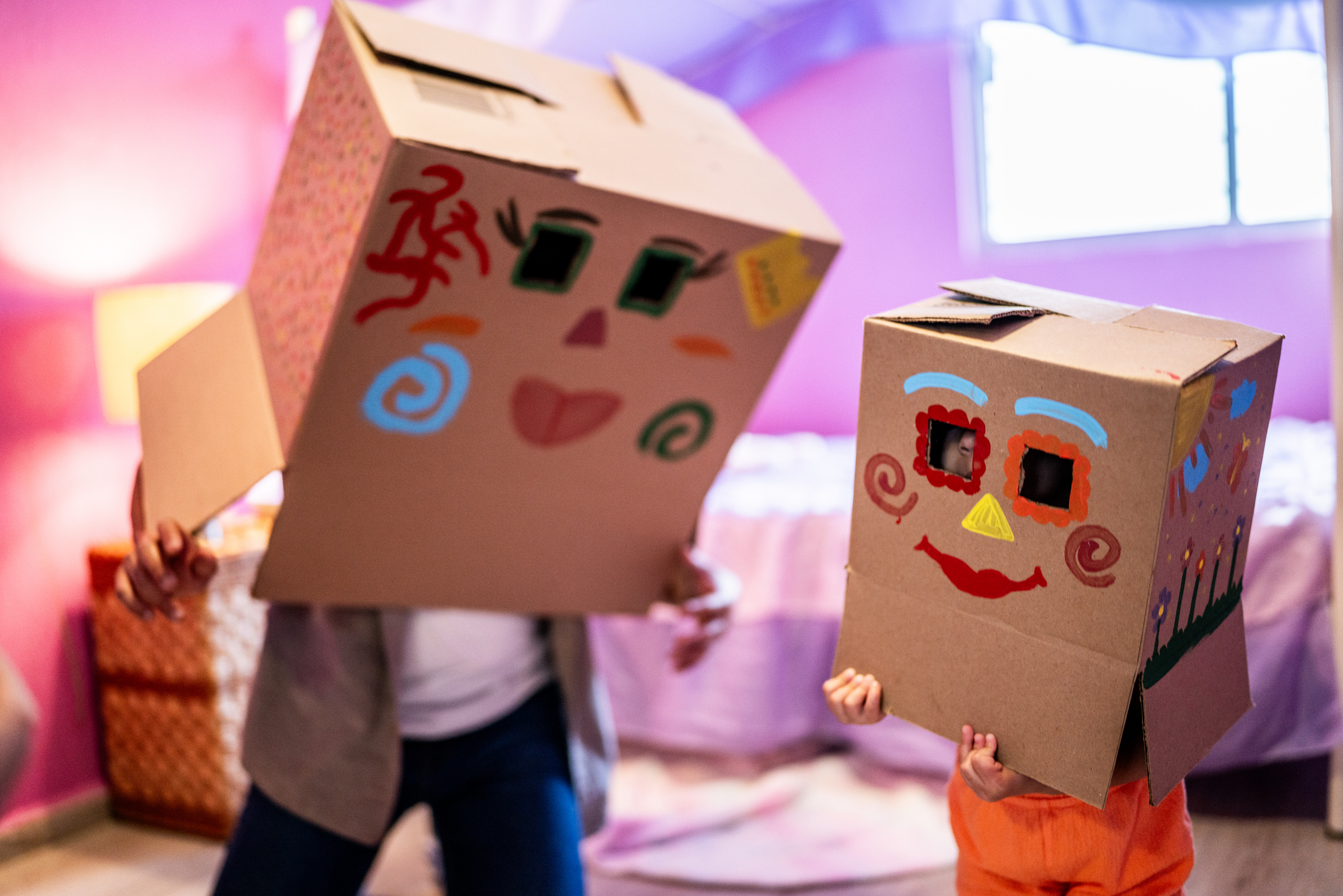 Two children wearing large cardboard boxes decorated with colourful painted faces, playing indoors in a pink room.
