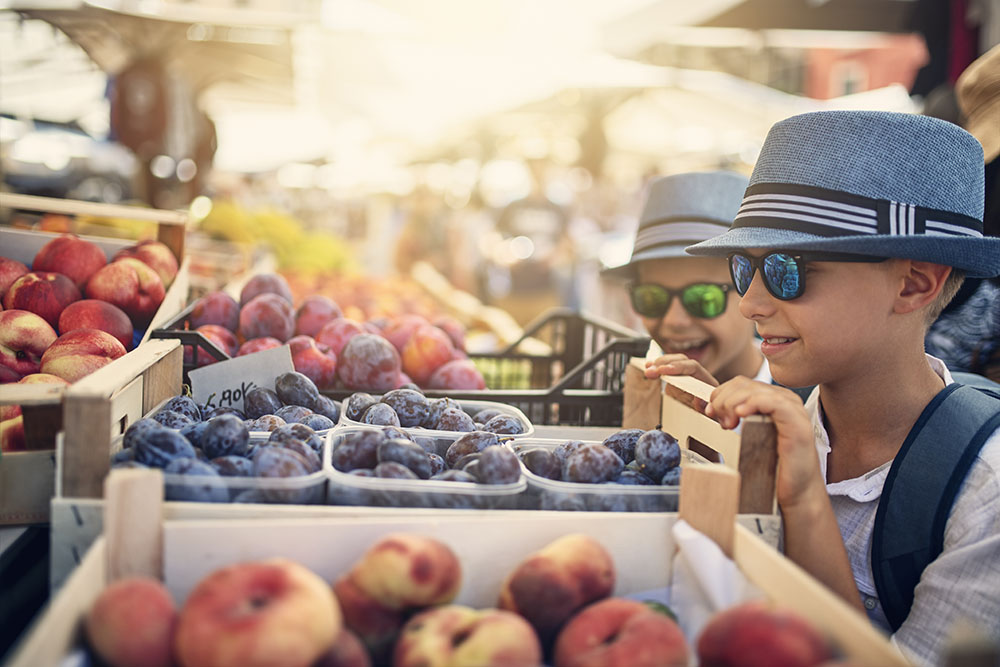 Two boys in hats and sunglasses look at trays of fresh fruit at an outdoor market.