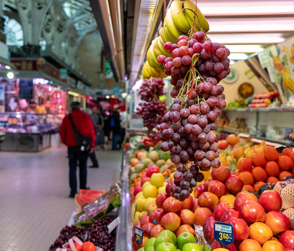 Fresh fruit including apples, grapes, and bananas displayed in a vibrant indoor market with shoppers in the background.
