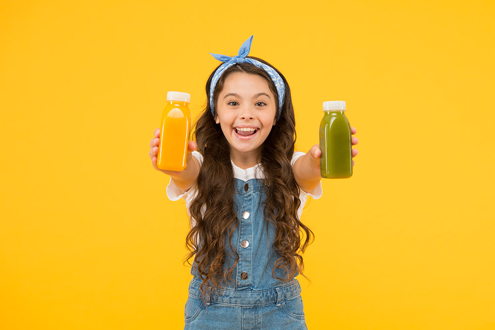 Child holding orange and green juice bottles, smiling against yellow background.