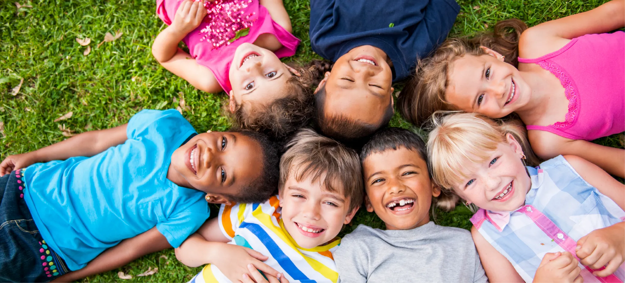 A group of smiling children from diverse backgrounds lying in a circle on grass, looking up.