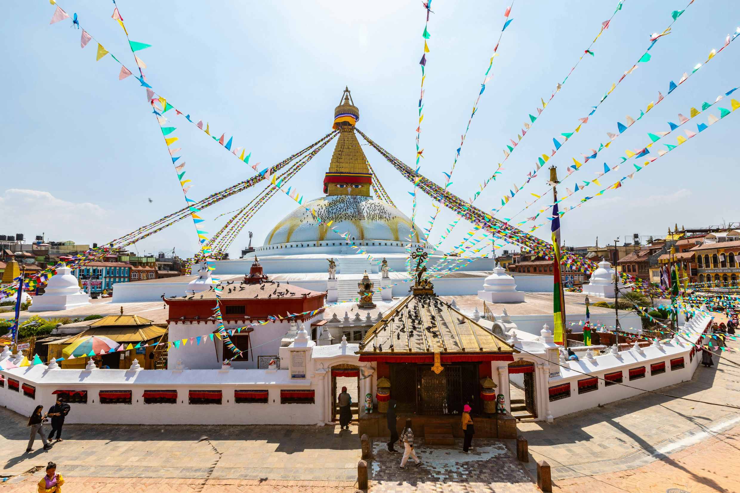Large Buddhist stupa with golden spire, colourful prayer flags, and people walking around the courtyard.