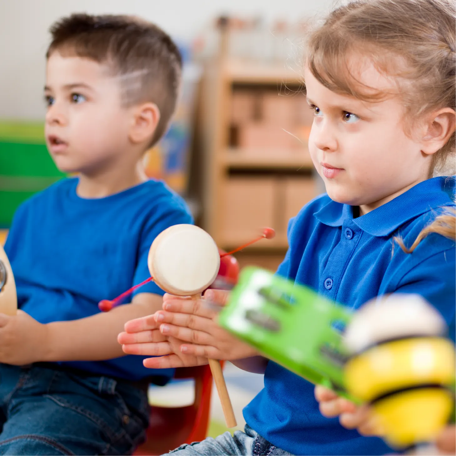 Two young children in blue shirts playing percussion instruments, including a tambourine and a hand drum, during a classroom music activity.