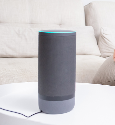 A child and an adult smiling while sitting on a sofa, looking at a smart speaker on a table.