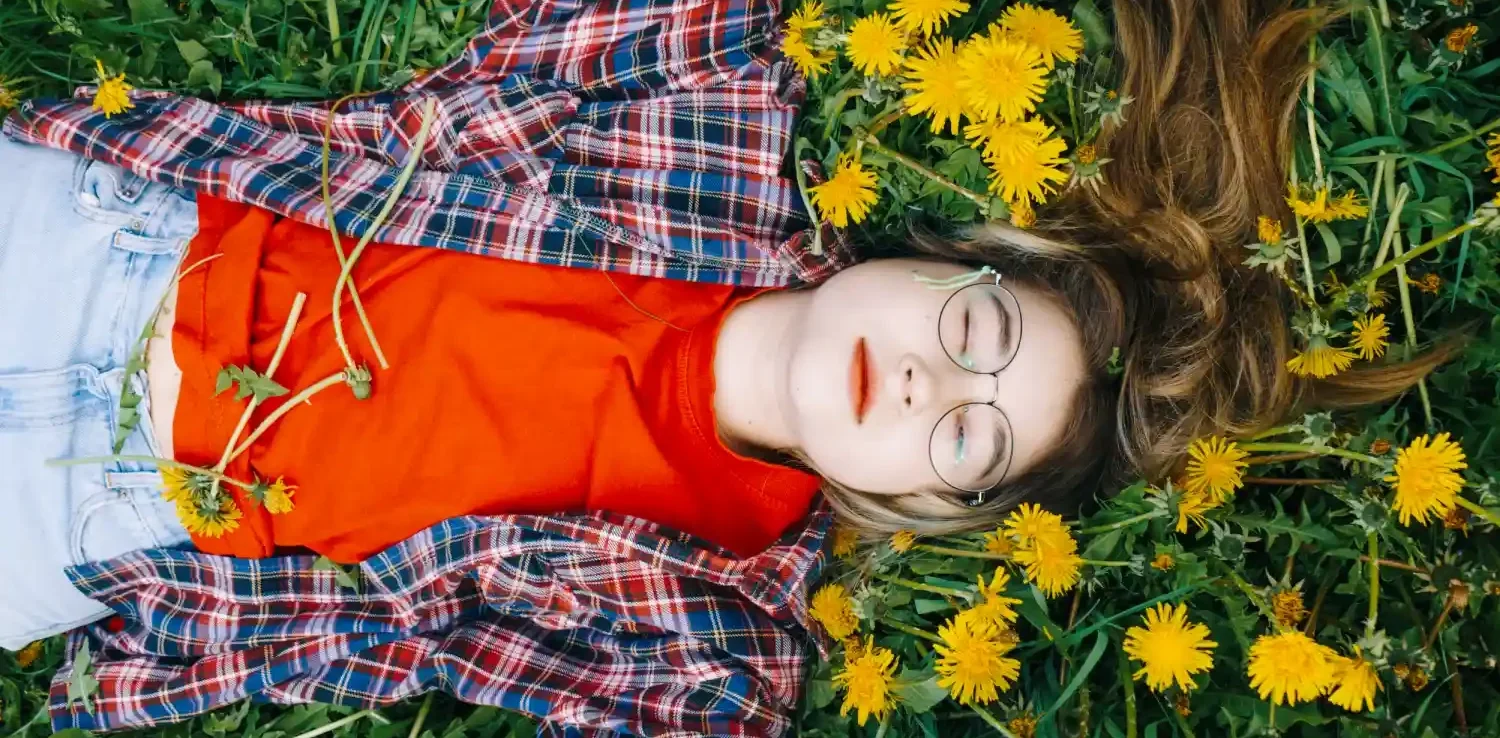 Child lying on back in a field of yellow dandelions, eyes closed, wearing glasses, red shirt, and plaid jacket, looking relaxed.