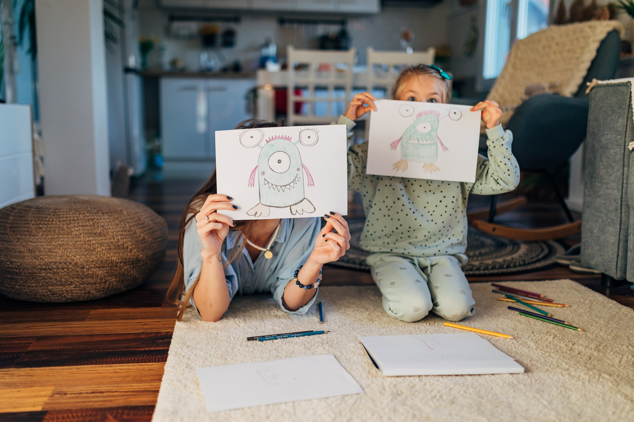 Child and adult holding monster drawings in front of their faces, sitting on the floor with coloured pencils and sketchbooks.