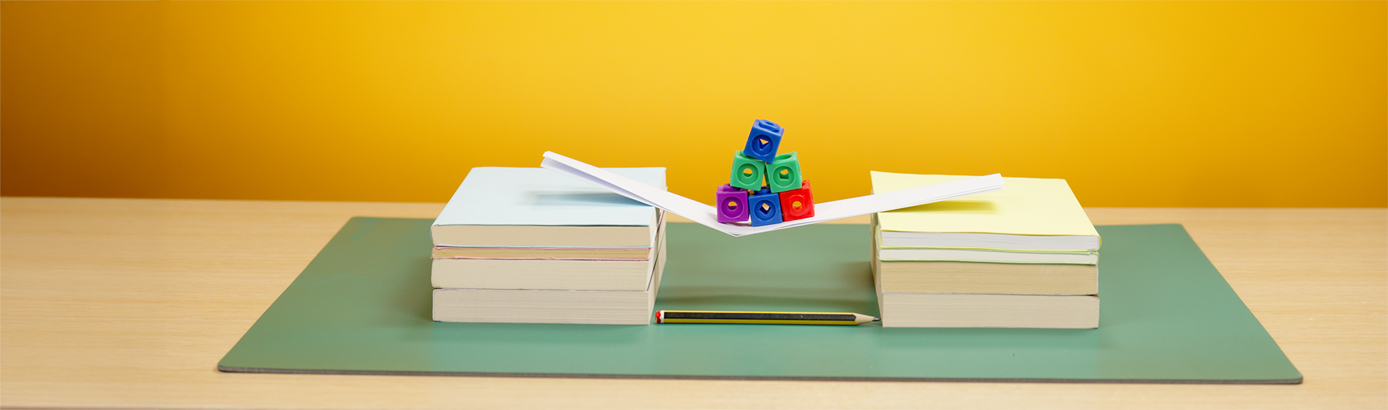 Two piles of books spaced around 15cm apart with a piece of paper folded twice and laid across the books like a bridge. There are six connecting cubes placed on the paper bridge and it is bending slightly.