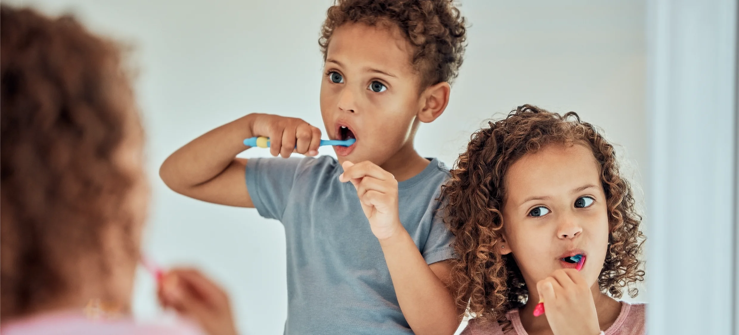 Two young children brushing their teeth infront of a mirror.