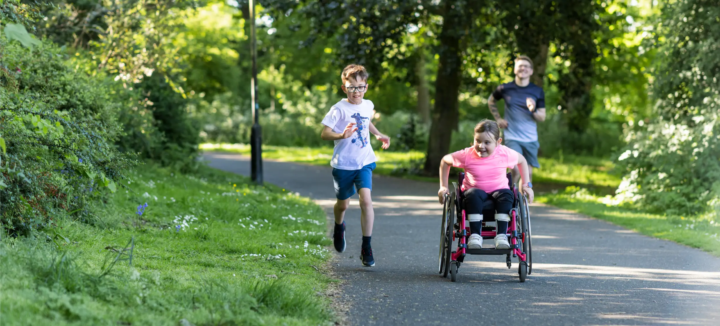 Two children race along a park path, one running and one using a wheelchair.