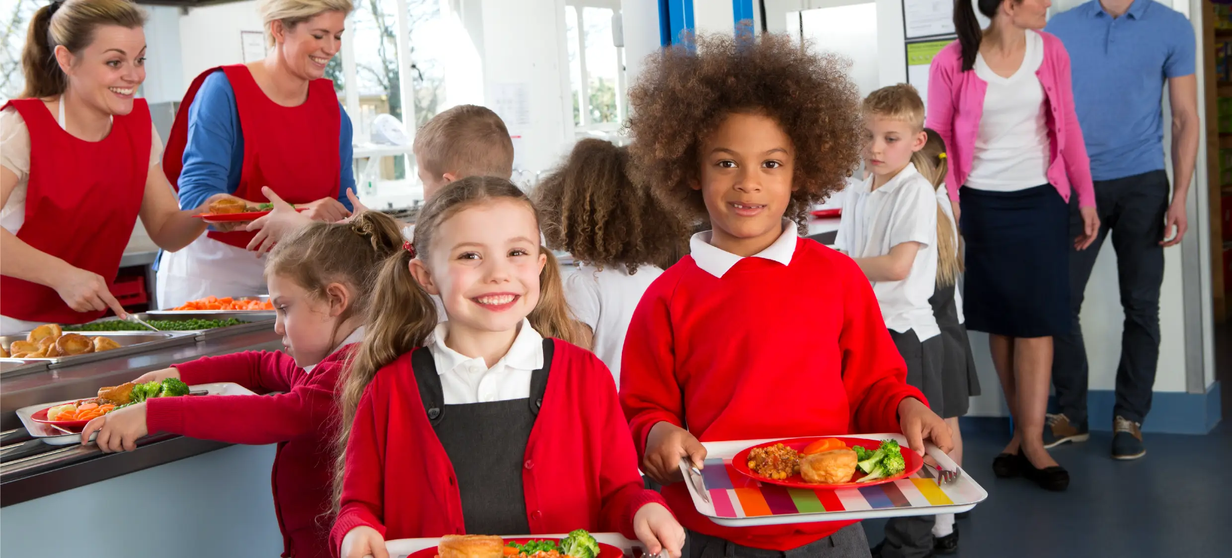 Children carry lunch trays while queuing for food in a school canteen.