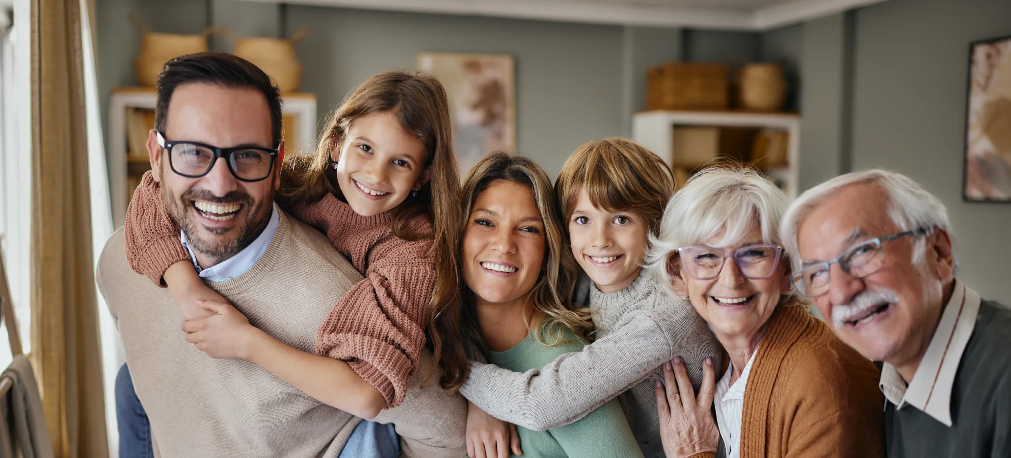 A multi-generation family smiling together indoors, with grandparents, parents and two children standing close.