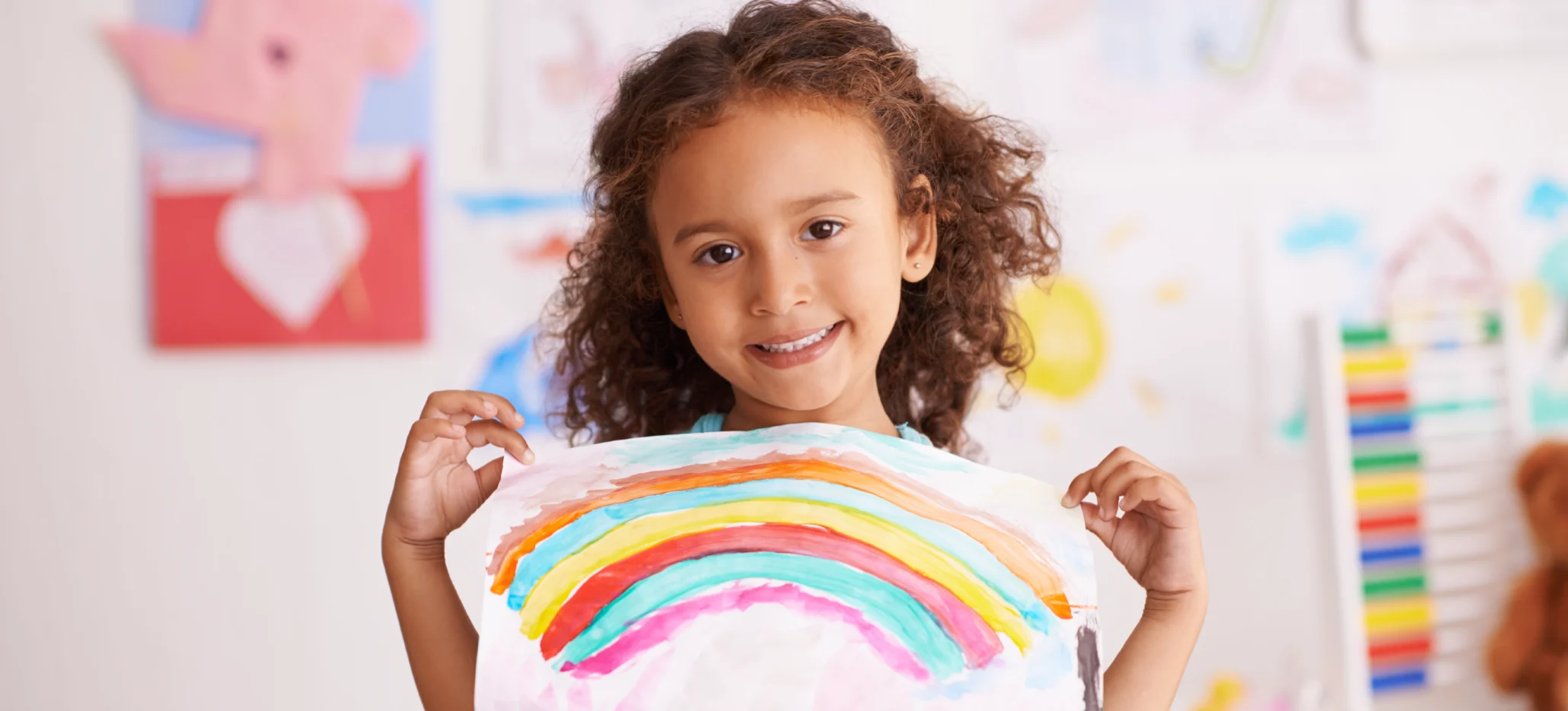 A smiling young child holding up a painting of a colourful rainbow.
