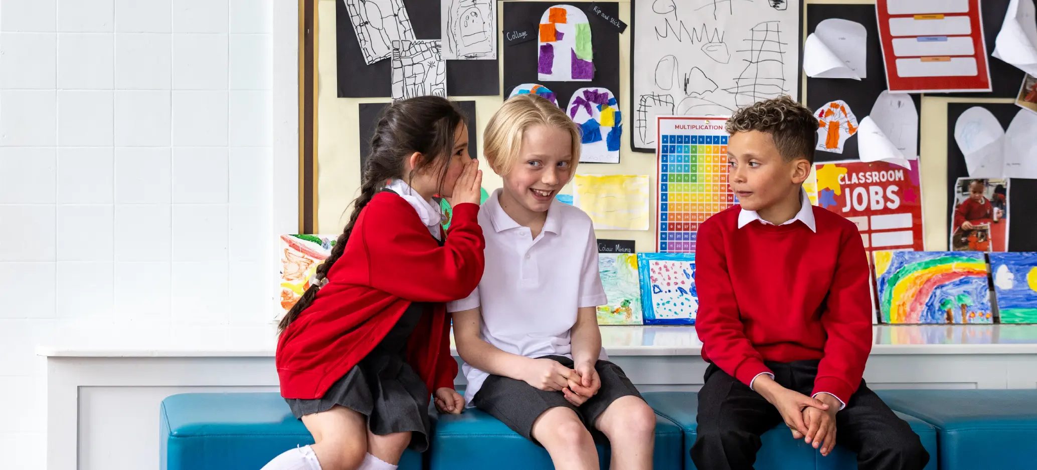 Three primary school children sitting together, with one child whispering to another while a third looks on.