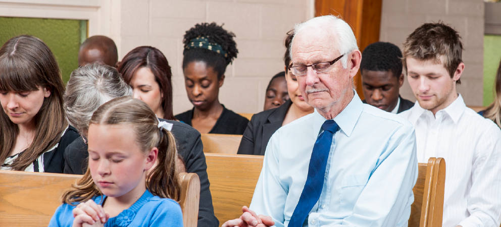 A group of people of different ages sitting in a church with their heads bowed and eyes closed, appearing to pray.