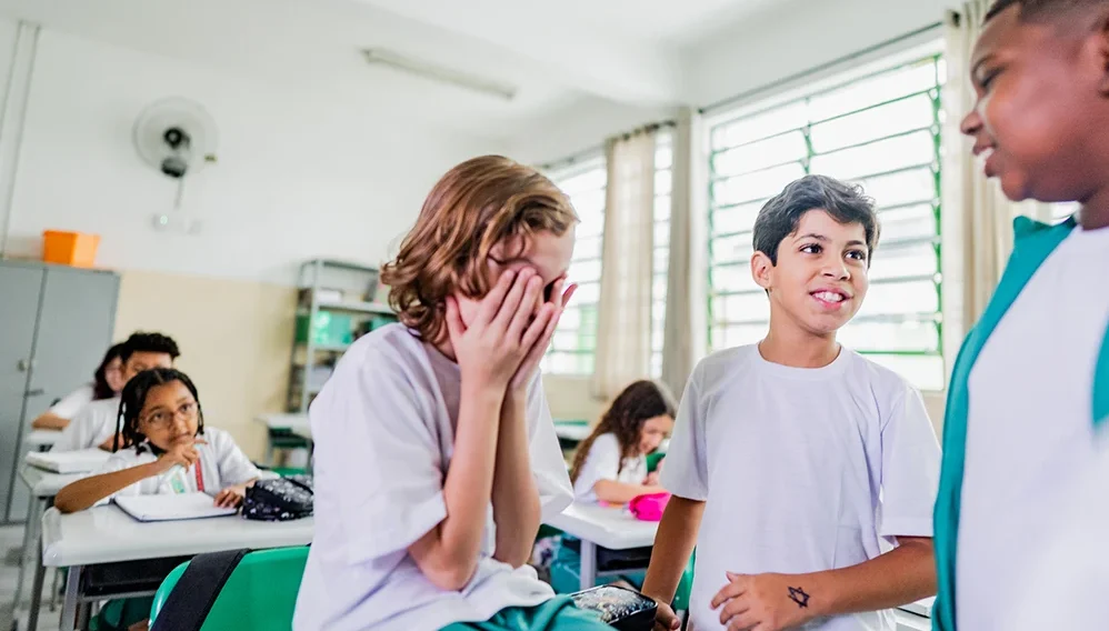 Pupils in a classroom, one child covering their face while others talk nearby.