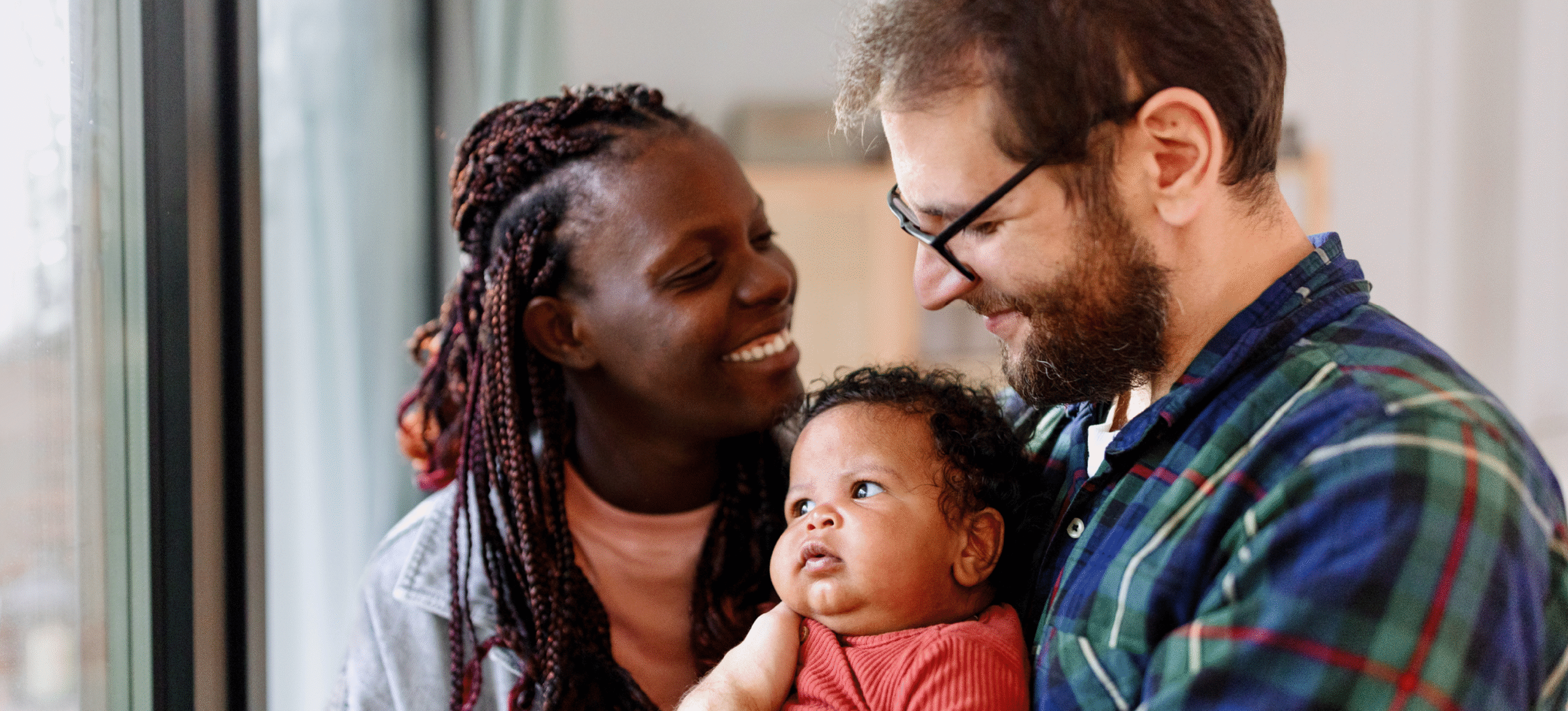 Two parents smiling at their baby while holding them at home.