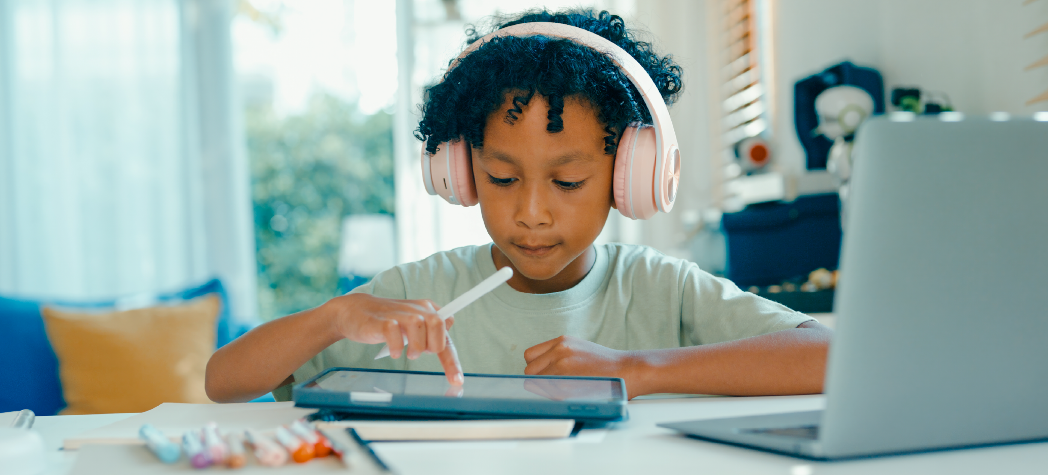 A child wearing headphones uses a tablet with a stylus and touchscreen while sitting at a table. A laptop is open nearby, and art supplies are spread out on the table.