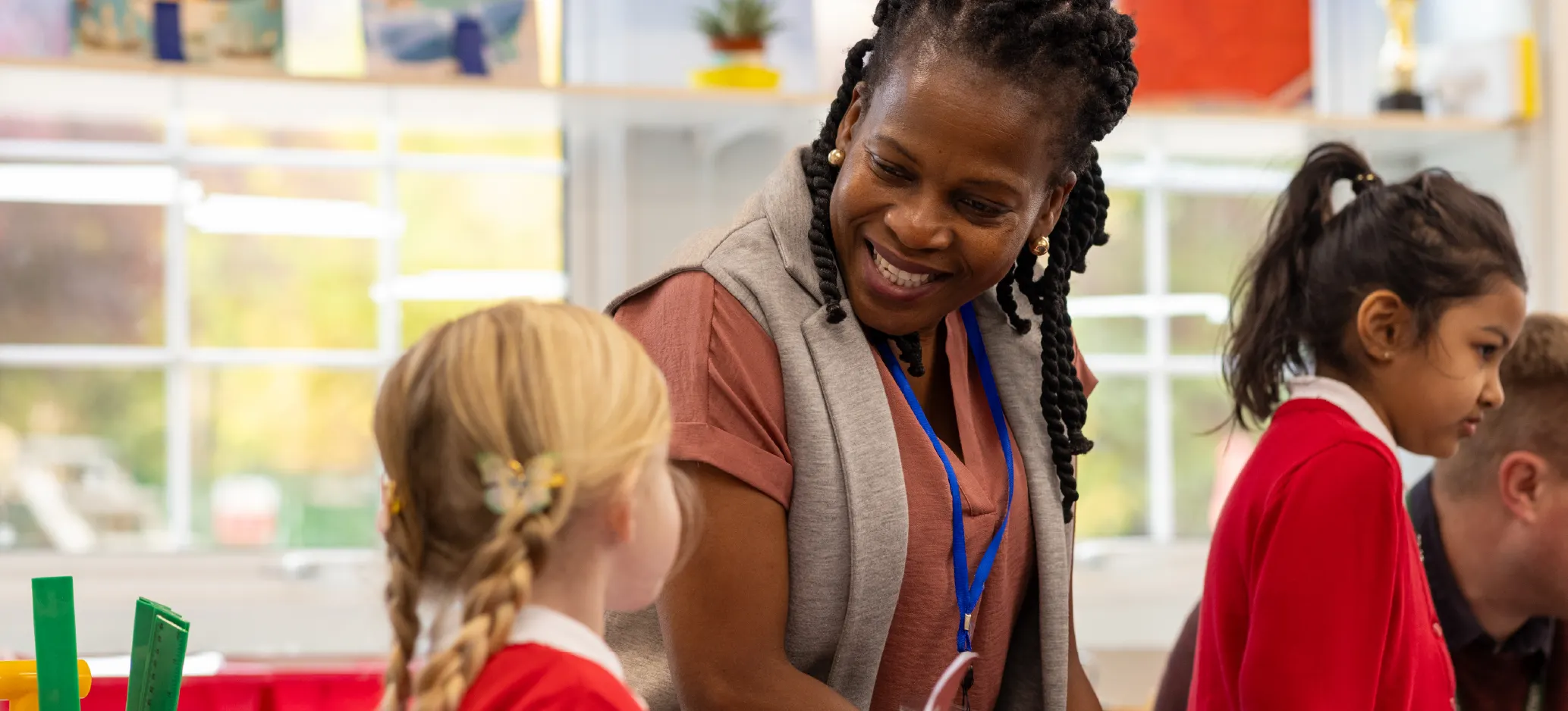 A teacher smiles and talks with a young pupil in a primary school classroom while other children work alongside them.