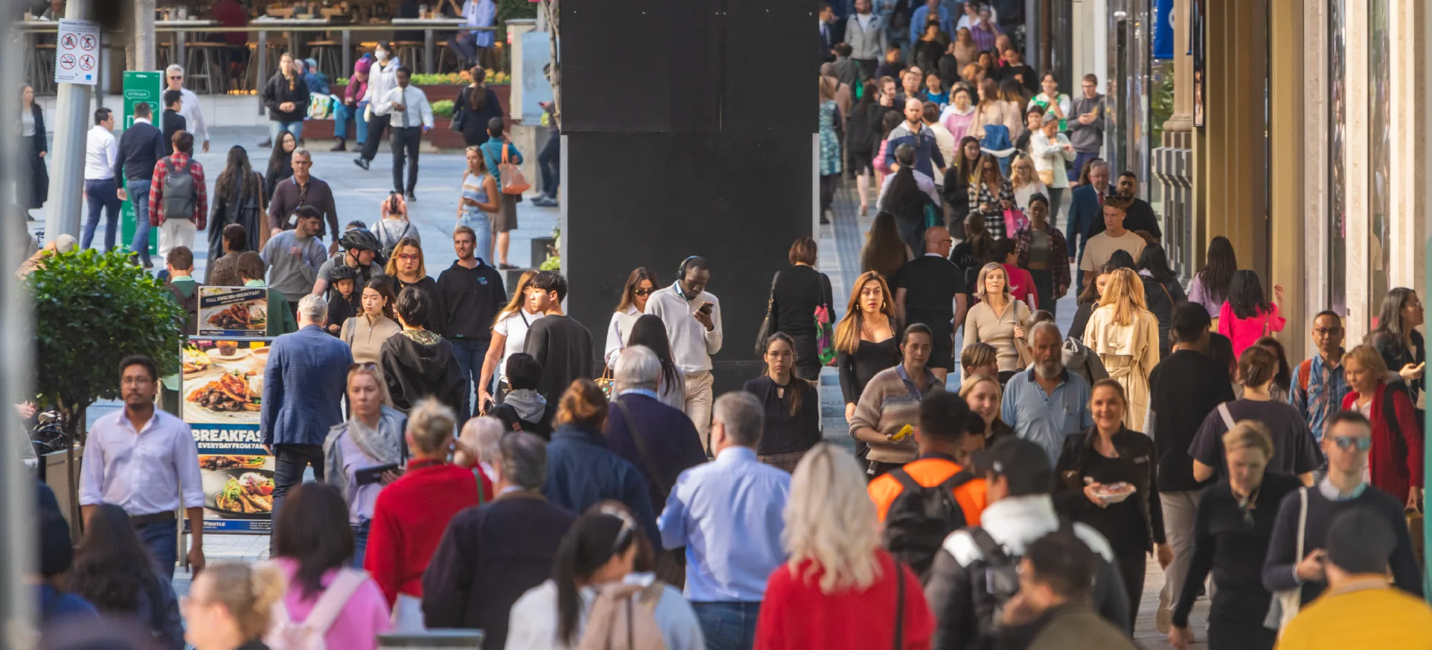 A large crowd of people walk along a busy city street lined with shops and restaurants.