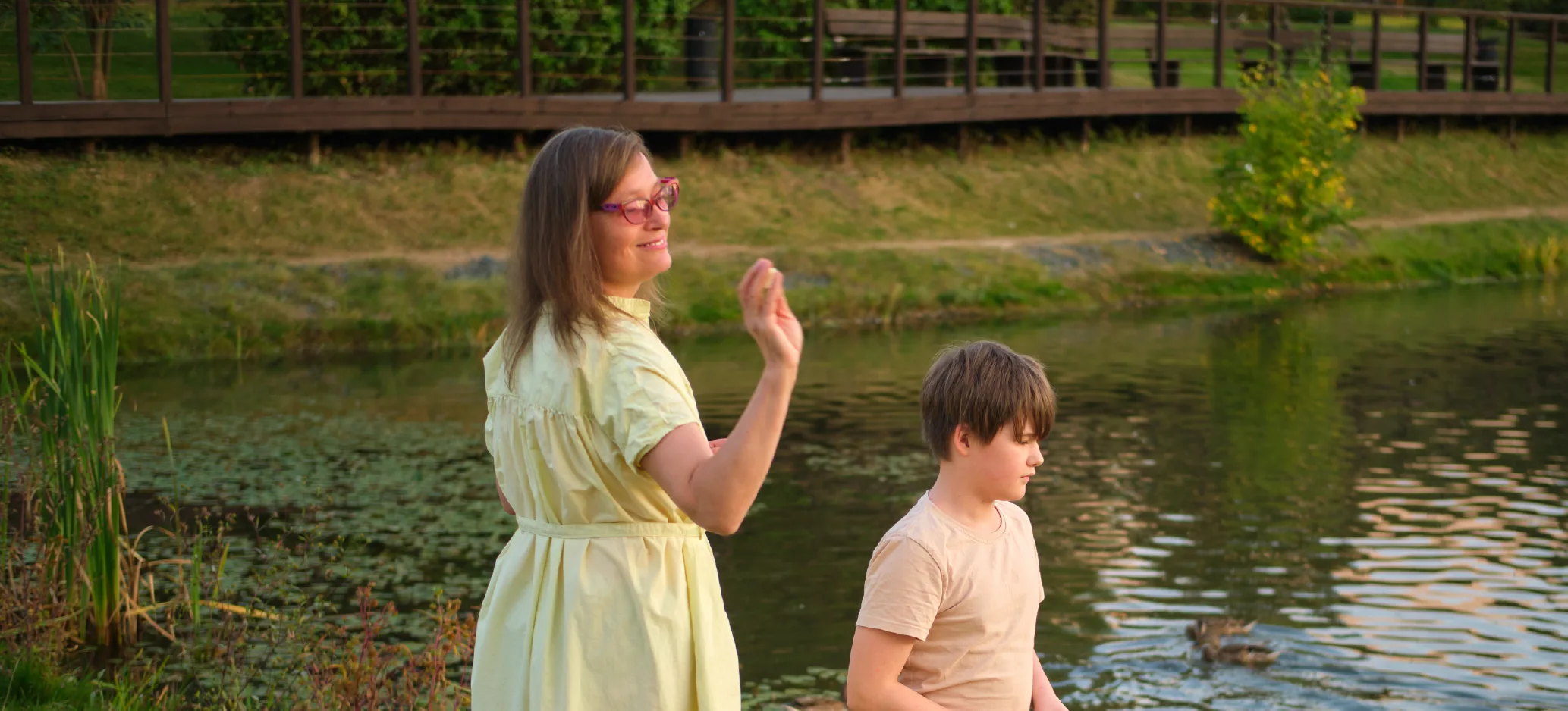 An adult and a child stand beside a pond in a park, watching ducks on the water while the adult waves.