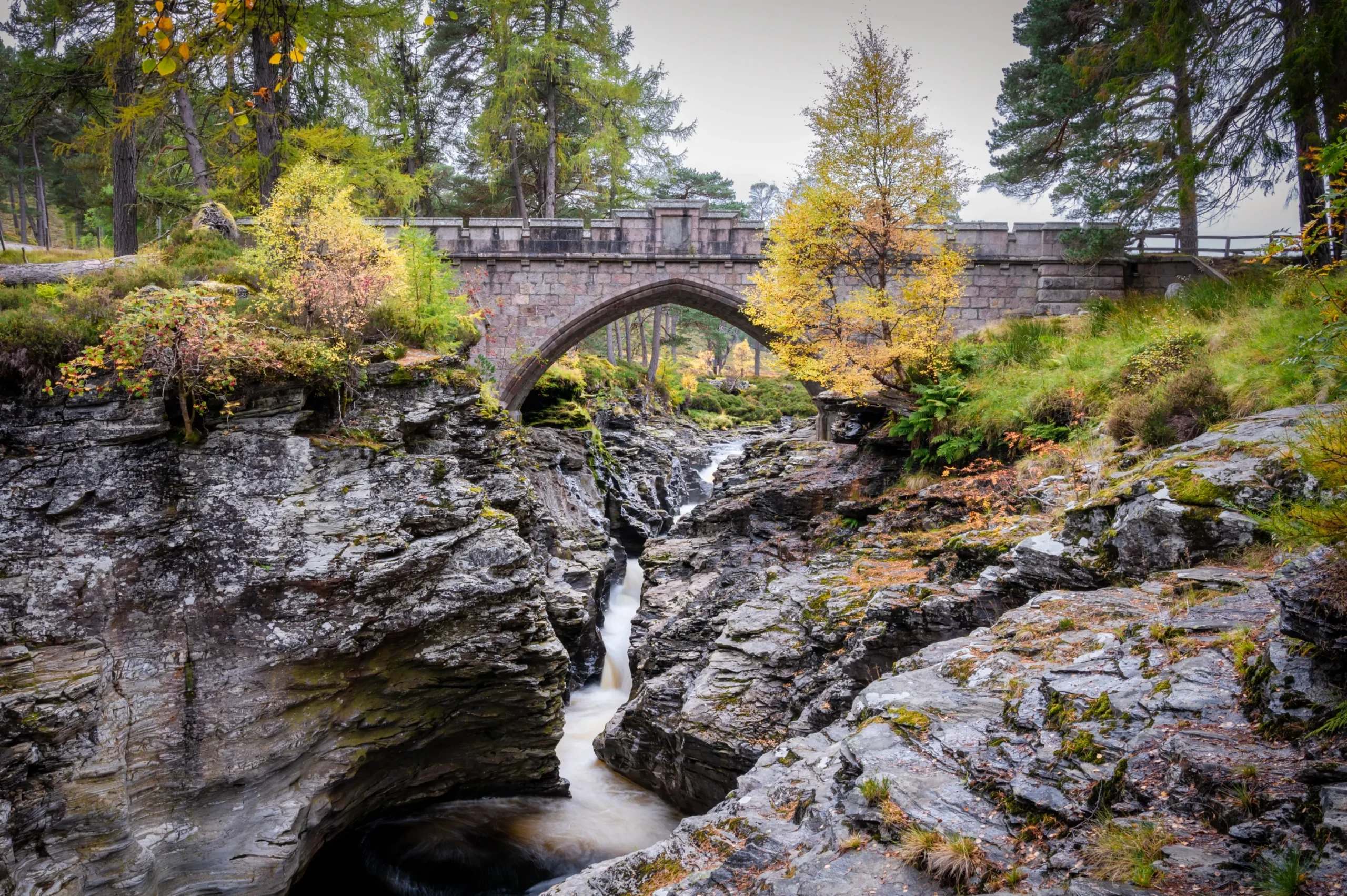 Stone arch bridge spanning a rocky river gorge surrounded by woodland and autumn trees.