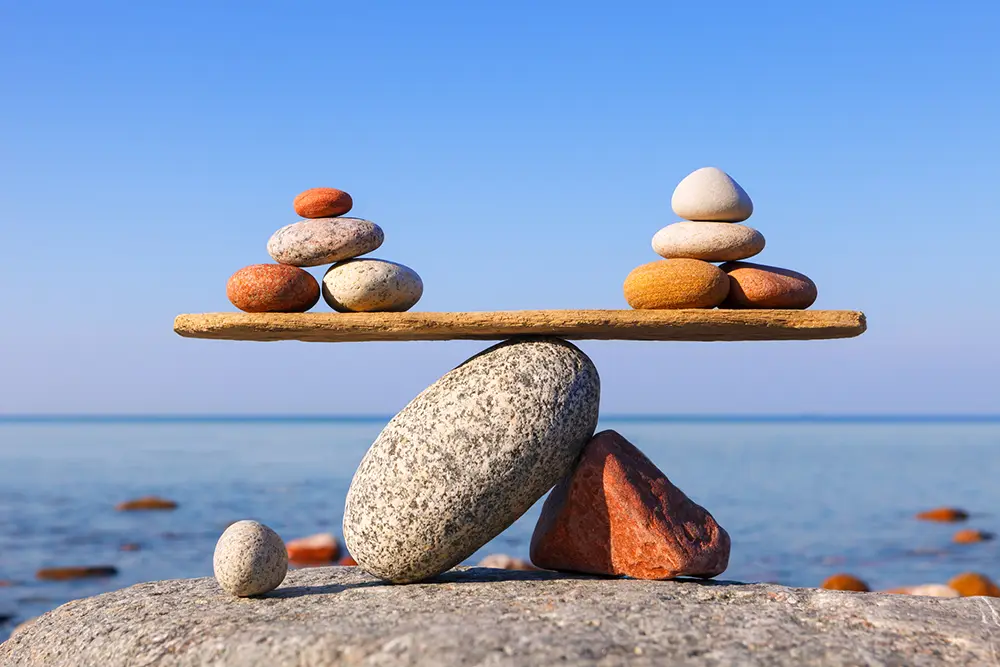 Two stacks of pebbles balanced on opposite ends of a wooden plank supported by rocks beside the sea.