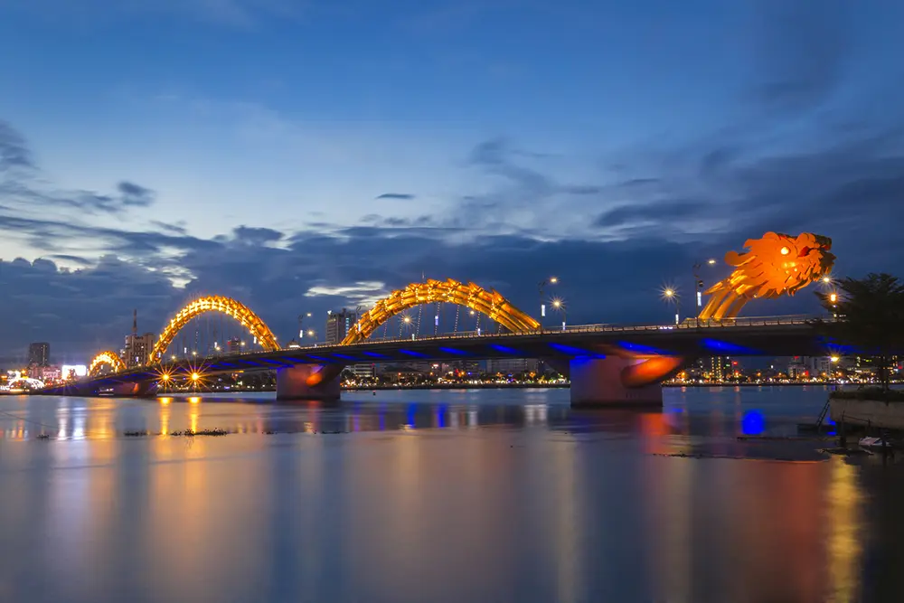 The Golden Dragon bridge in Vietnam at dusk. The bridge is lit up red and orange.
