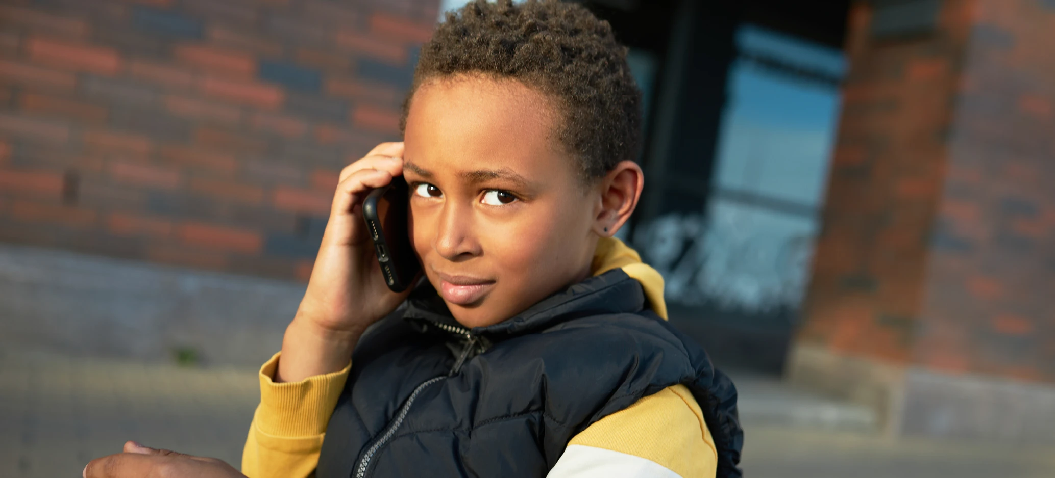 A young boy wearing a yellow hoodie and navy padded jacket holds a mobile phone to his ear while standing outdoors.