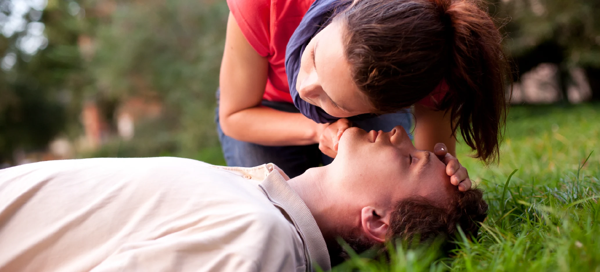 A woman checks an unconscious man's breathing by placing her ear close to his mouth while he lies on the grass.