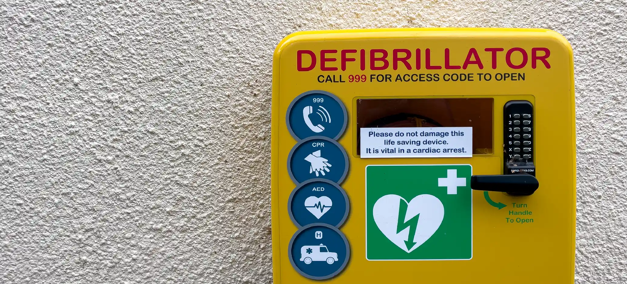 A yellow public access defibrillator cabinet mounted on an outdoor wall, with a keypad, handle and instructions to call 999 for an access code.