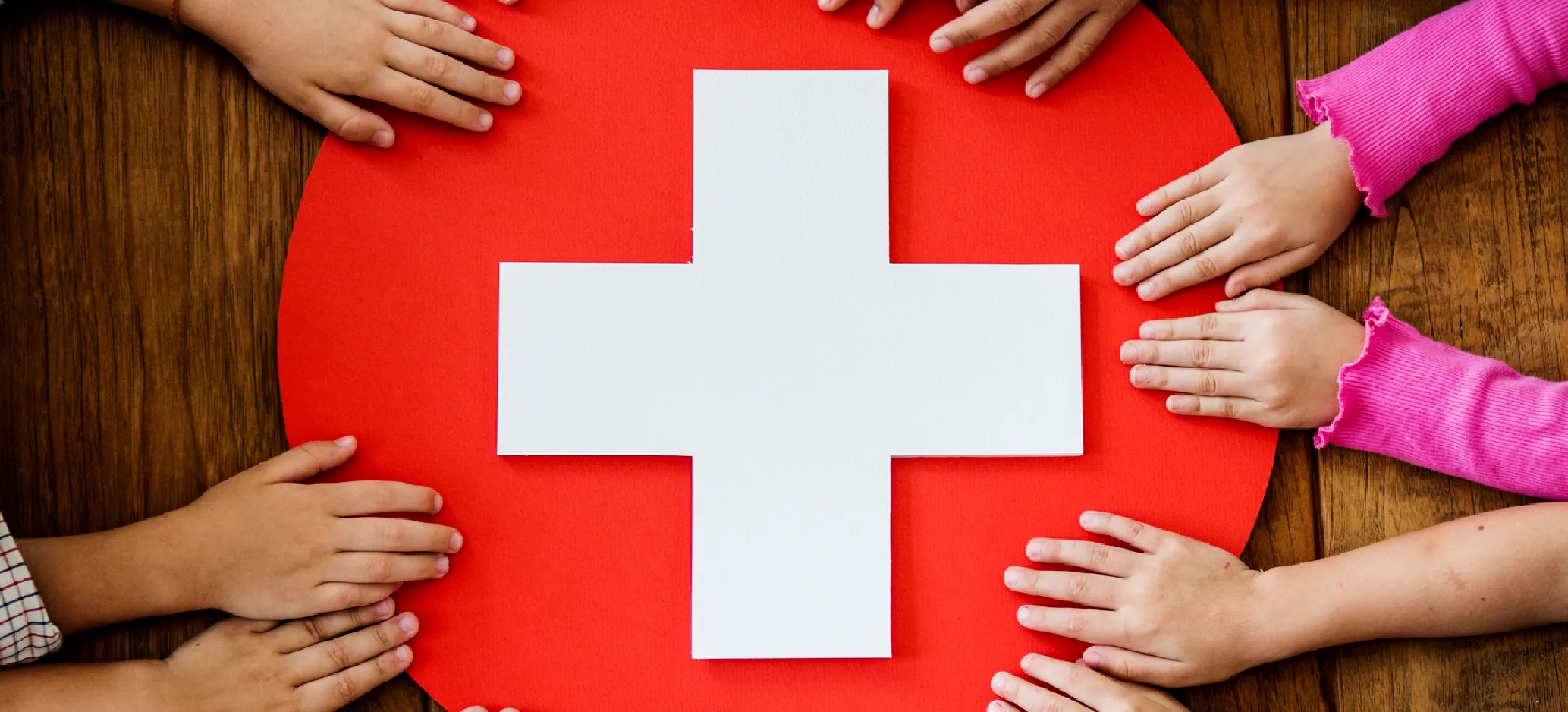 Children's hands surround a large white first aid cross on a red circular background, placed on a wooden table.