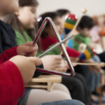 Image of a child's hand holding a triangle with other children faded in the background holding other instruments during a music lesson
