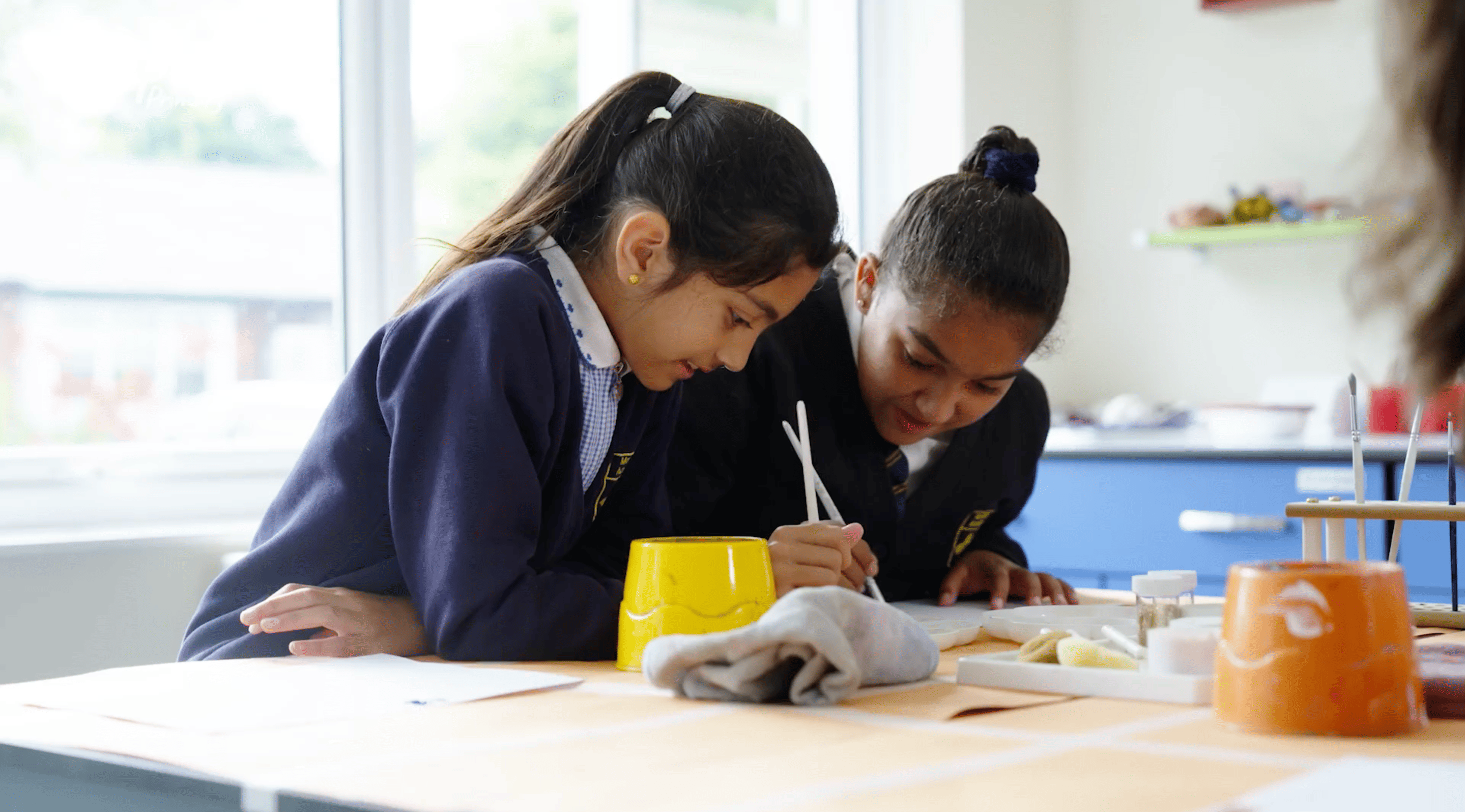 Two primary school pupils in navy blue uniforms are engaged in an art activity, carefully painting on paper. They are smiling and focused, using fine brushes and a palette of paints. The table is covered with art supplies, including water pots, paintbrush holders, and sponges. The background features a bright classroom with large windows and blue storage units.