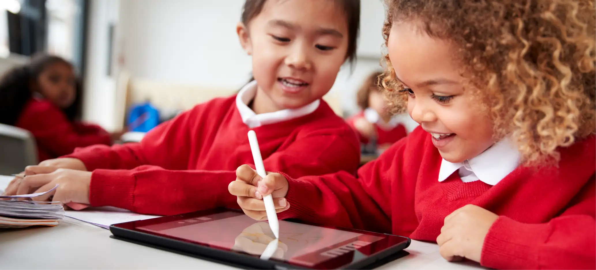 Two primary pupils in red uniforms happily using a tablet together in class.