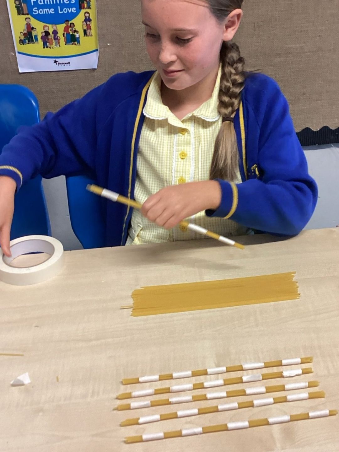 Pupil in blue uniform wrapping tape around spaghetti sticks for a model project.
