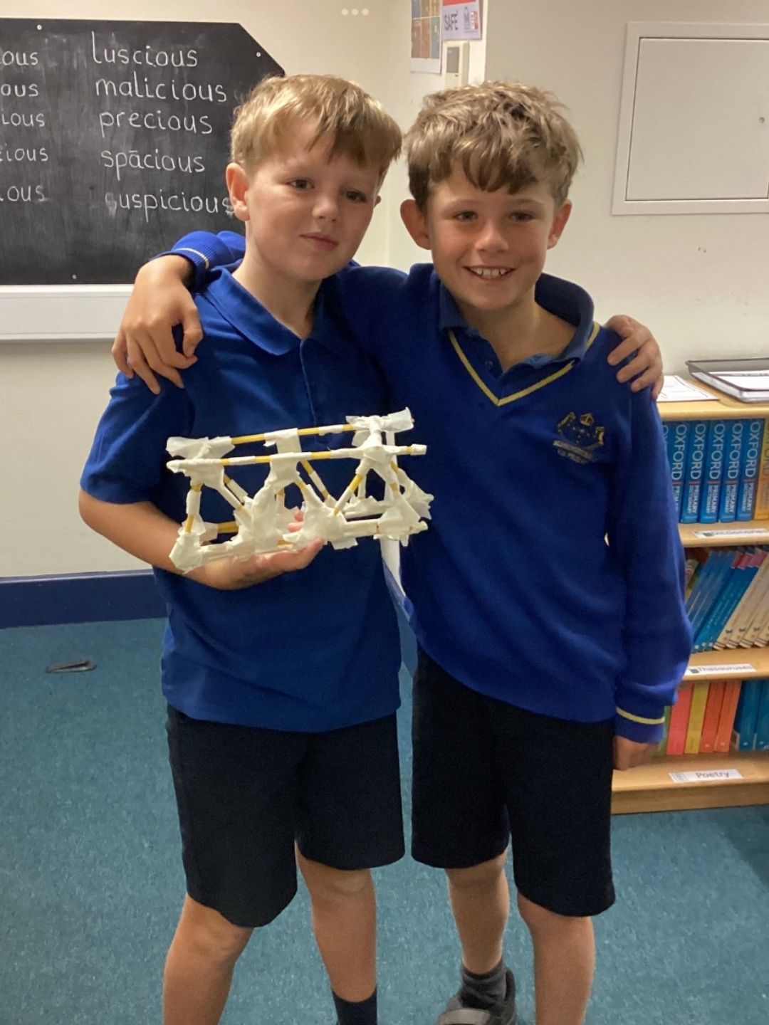 Two pupils in blue uniforms holding a model structure made from straws and tape.