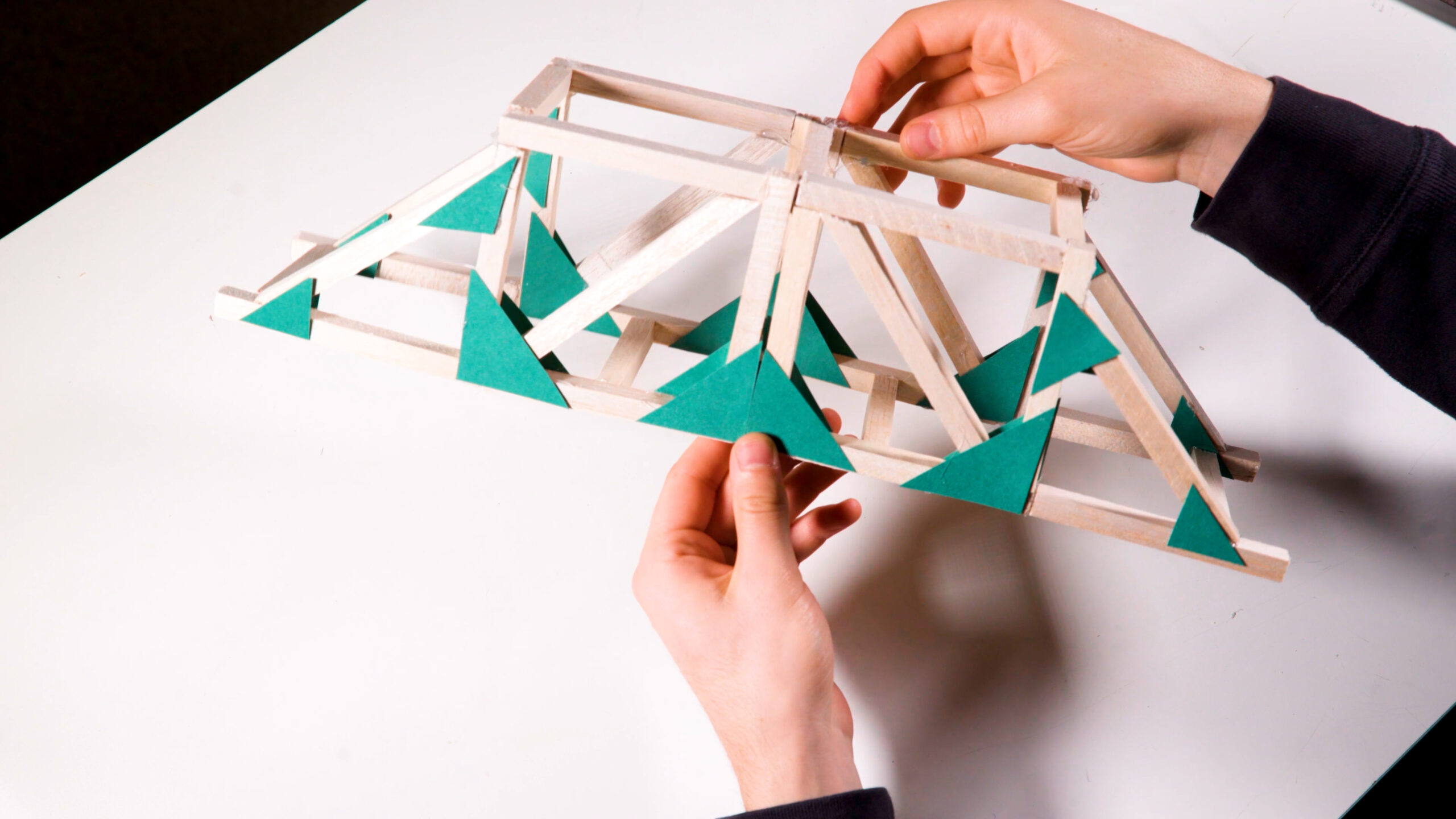 Hands holding a small model bridge made from wooden sticks and green triangles.