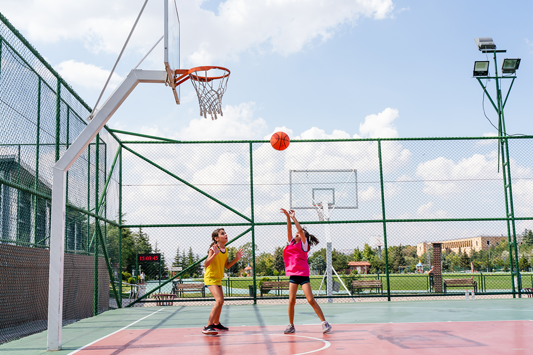 Two girls playing basketball on an outdoor court, one shooting the ball while the other defends.
