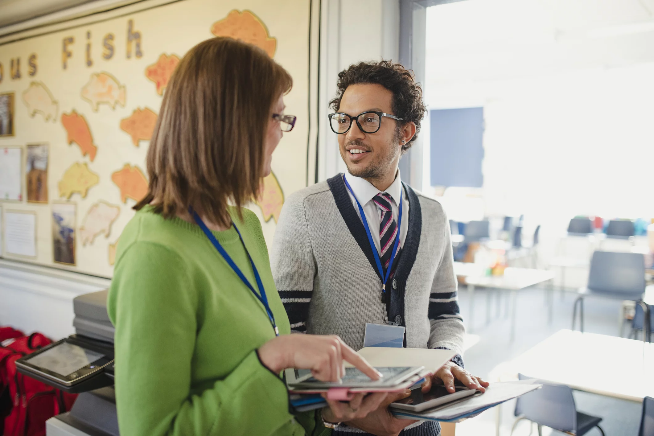 A mature woman discusses her work with her colleague; they are both carrying digital tablets and paper work. This is a school in Hexham, Northumberland in north eastern England.