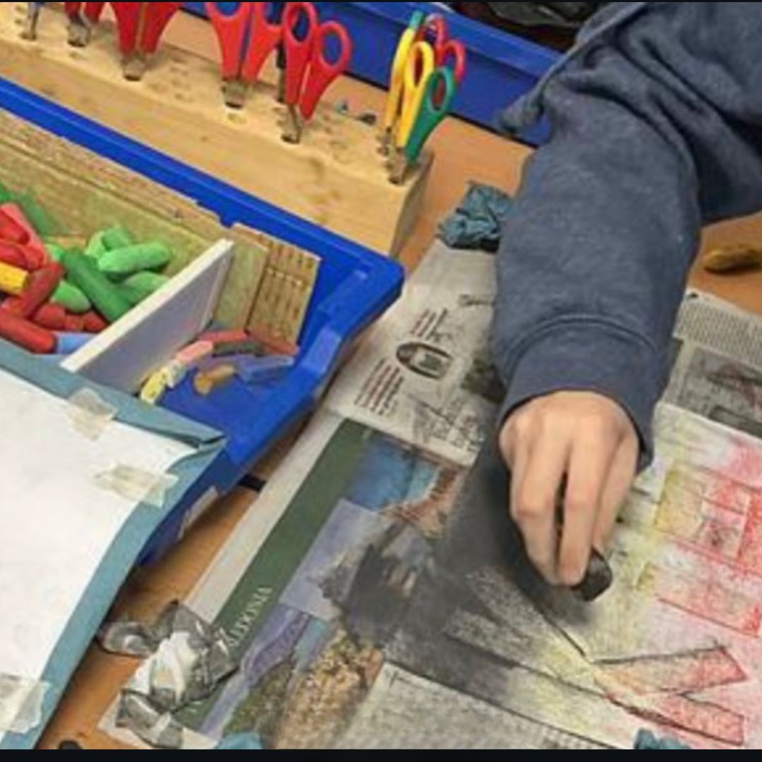 Child shading a drawing with charcoal on newspaper covered table.