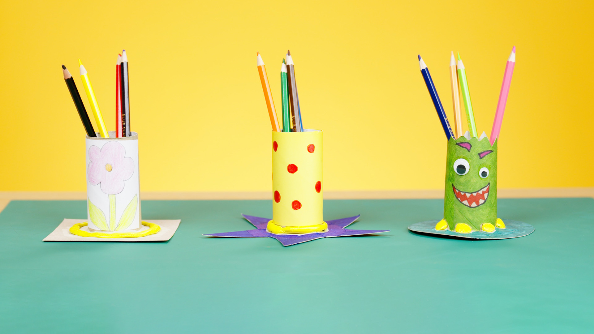 Three pencil pots made from cardboard tubes with different designs to appeal to a Year 1 pupil, on a yellow background.