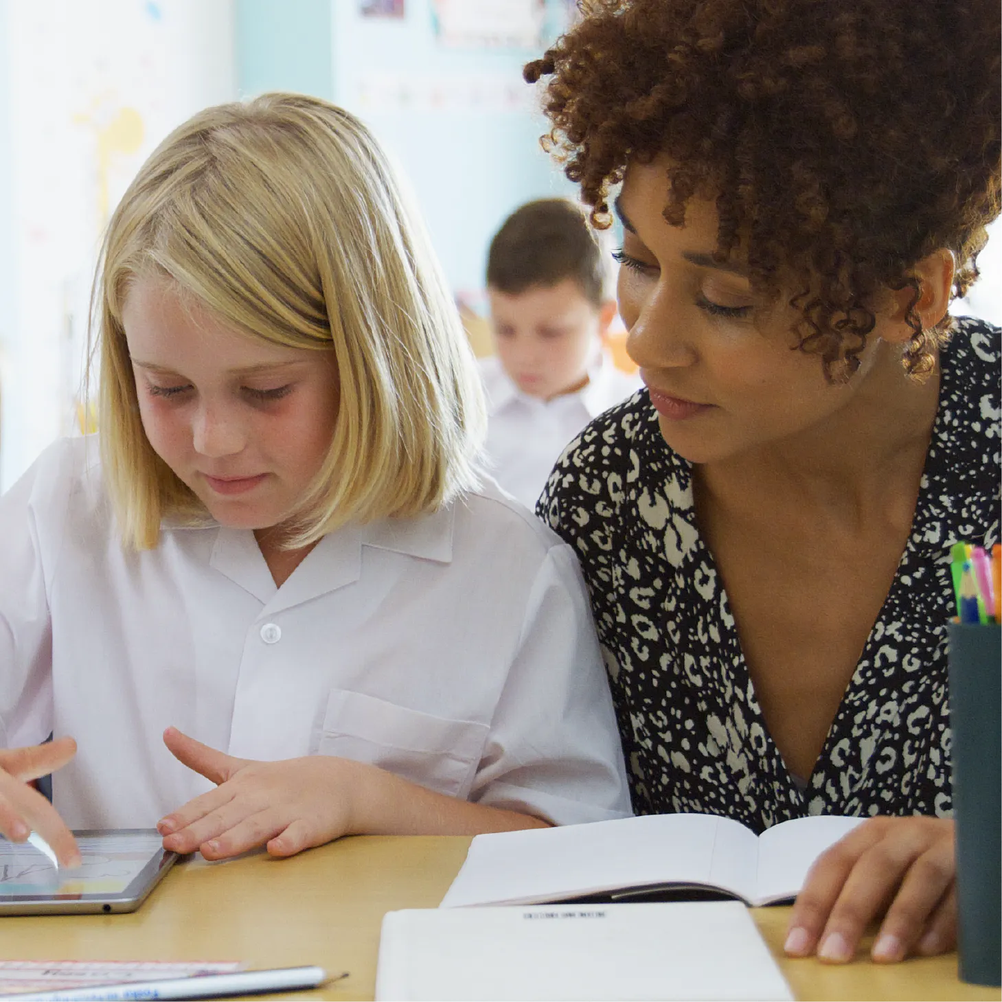 A teacher observing a young pupil in class while she navigates an app an on electronic tablet.
