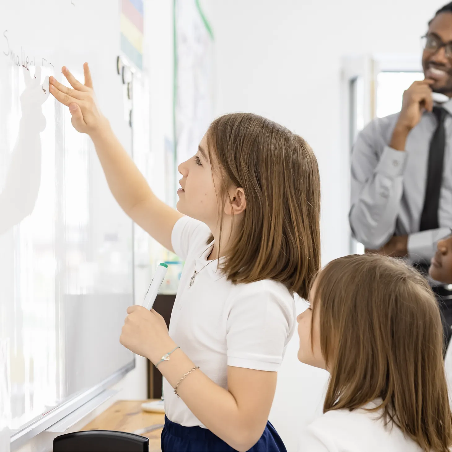A young girl is rubbing out a mistake on the class whiteboard while the teacher and a peer watch.