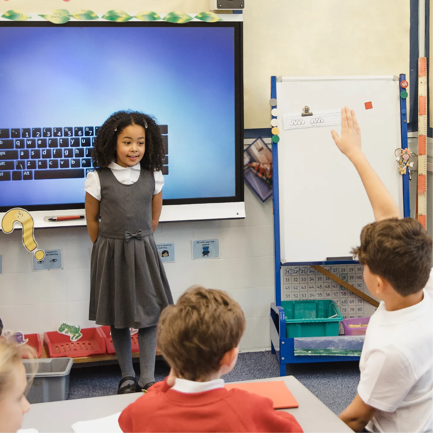 A young pupil talking to the rest of her class, with her peers holding up their hand.