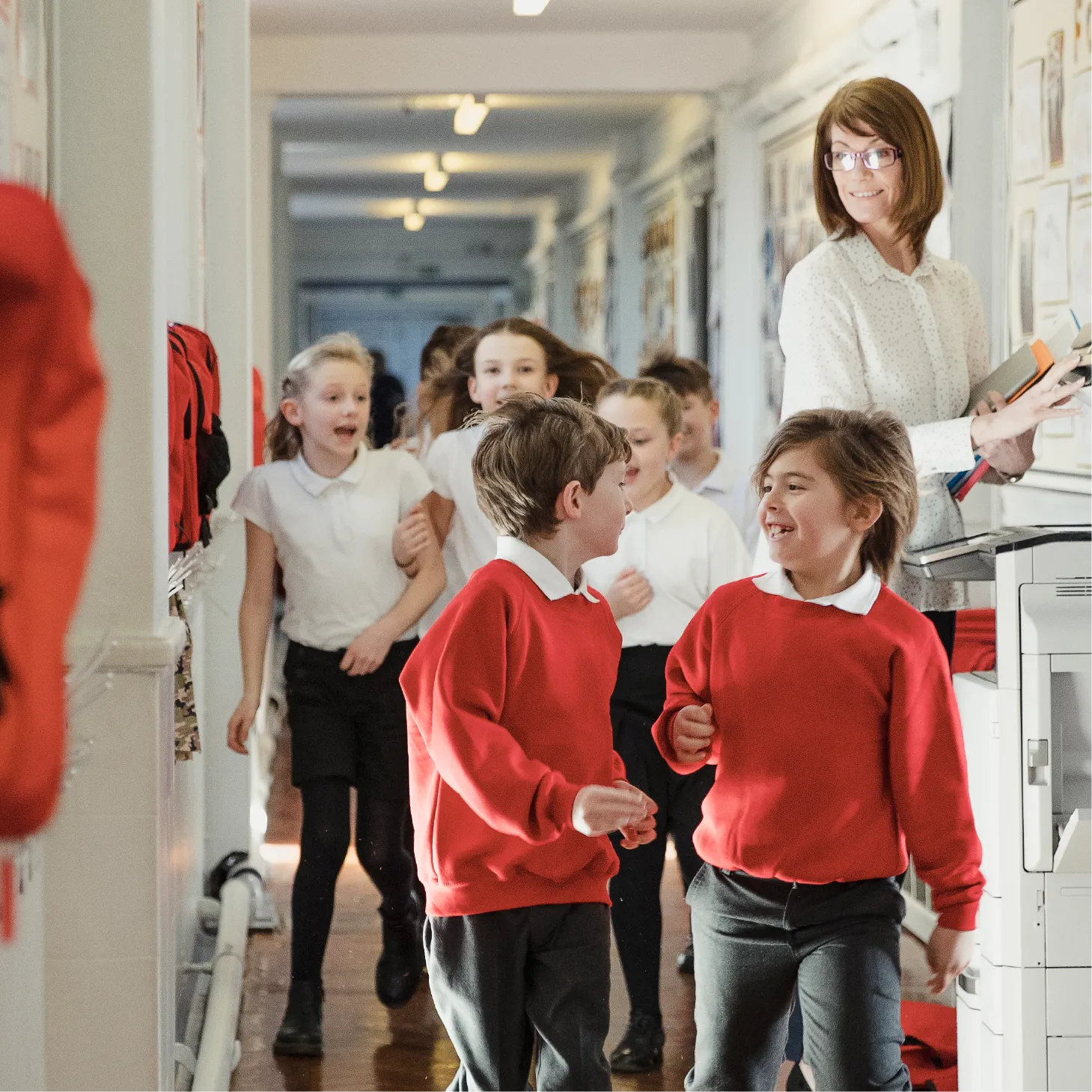 Children walking down a school corridor, while a teacher uses a photocopier and keeps watch.